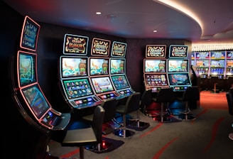 A row of brightly lit slot machines in a casino setting, each with colorful displays and various game themes. The machines are lined up against a dark wall and are unoccupied, with cushioned chairs placed in front of them. The area is dimly lit except for the glow from the screens, creating a vibrant and engaging atmosphere.